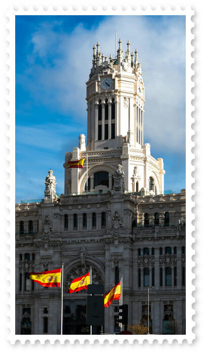 Palacio de Cibeles in Madrid with 3 Spanish flags in front and the blue-cloudy sky